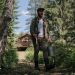 A man walks through a forest holding a fishing rod and bucket on a summer day.