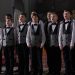 Group of boys singing in a choir, wearing formal uniforms in an indoor setting.